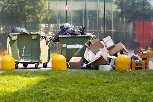 Two large green wheelie bins positioned on a paved area adjacent to a glass-fronted building, with one bin lids open and filled with black plastic rubbish sacks, and the other bin appearing relatively empty. To the right of the bins, there is a pile of flattened cardboard boxes, some partly torn, with visible corrugated texture in beige and brown shades. A small plastic crate containing miscellaneous waste items, including plastic bottles and packaging, is also present among the cardboard. Bright yellow concrete bollards are placed along the edge of the paved area, separating it from a well-maintained grassy verge in the foreground. Reflections of surrounding trees and vehicles can be seen in the building’s glass facade. The scene suggests a typical rubbish collection or clearance site outside an office or commercial property in Sidcup, where waste and recyclable materials are temporarily stored prior to collection by a professional waste management company such as Office Clearance Sidcup, serving the local postcode area.