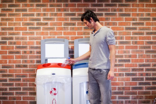 A young man with dark hair, wearing a light grey long-sleeve shirt and grey trousers, stands outdoors next to two white rubbish bins positioned against a red brick wall. The waste containers are made of plastic with integrated handles and decorated with red and blue recycling symbols, indicating their use for waste separation. The man is holding a small plastic water bottle and appears to be in the process of discarding it into the bin with the red lid. The setting suggests a residential or commercial environment, possibly near a property in Sidcup, Kent, with natural daylight illuminating the scene. The overall context relates to rubbish disposal and waste management services provided by Office Clearance Sidcup, emphasizing recycling efforts typical in local rubbish removal operations.