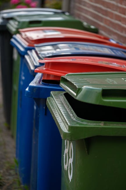 A worker wearing a blue uniform and an orange high-visibility vest is depicted emptying a blue recycling bin into the open rear compartment of a large white waste collection truck. The truck, position