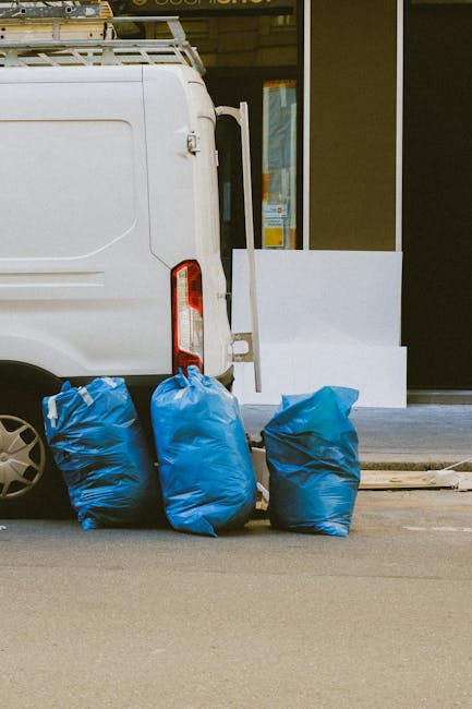 A white commercial van is parked on a street, with three large blue rubbish bags placed on the pavement near the rear end of the vehicle, likely prepared for collection or disposal. The bags are made