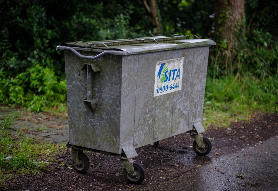 A row of four white plastic wheelie bins with closed lids, situated on an asphalt surface in front of a tall, dark-looking shipping container. The bins are positioned side by side, each with small whe